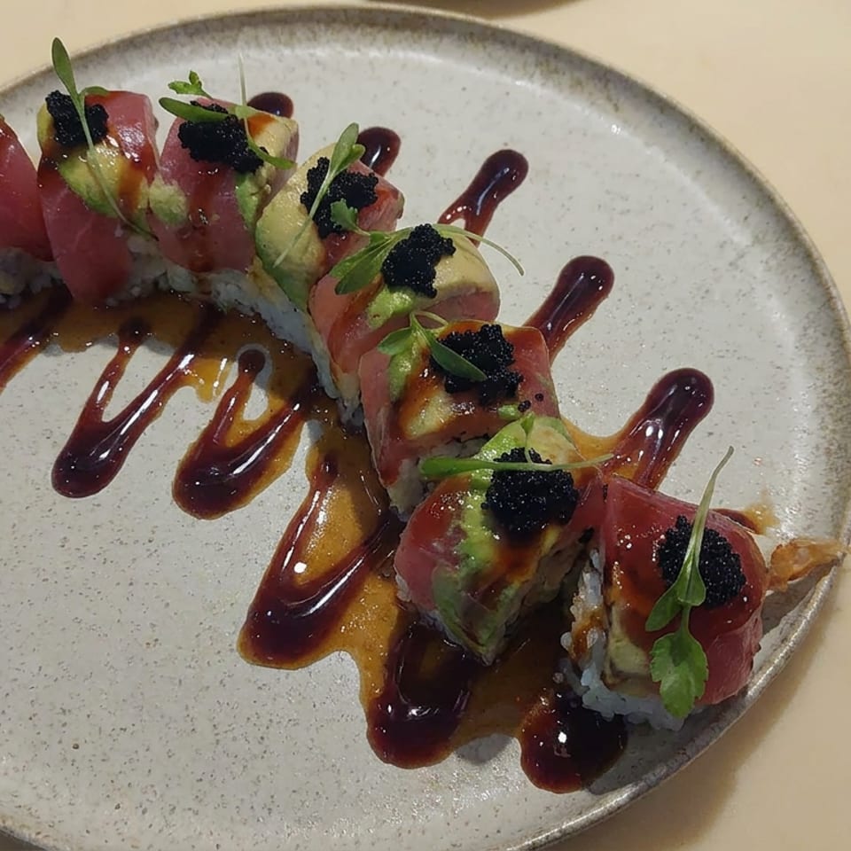 Sushi roll pieces topped with tuna, avocado, black roe, microgreens, and drizzled sauces on a ceramic plate.