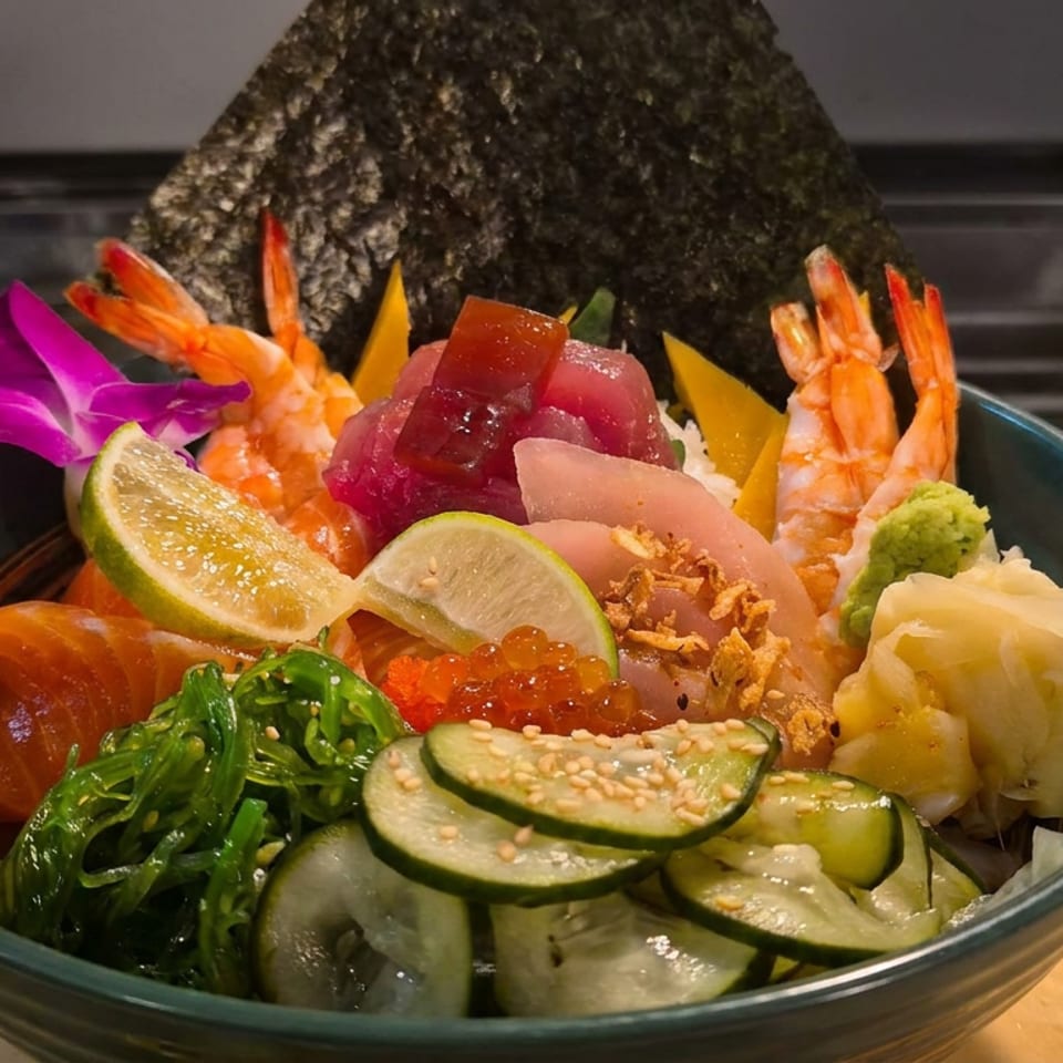 Colorful seafood poke bowl with shrimp, tuna, salmon roe, vegetables, lime, seaweed, dumplings, and a nori sheet.